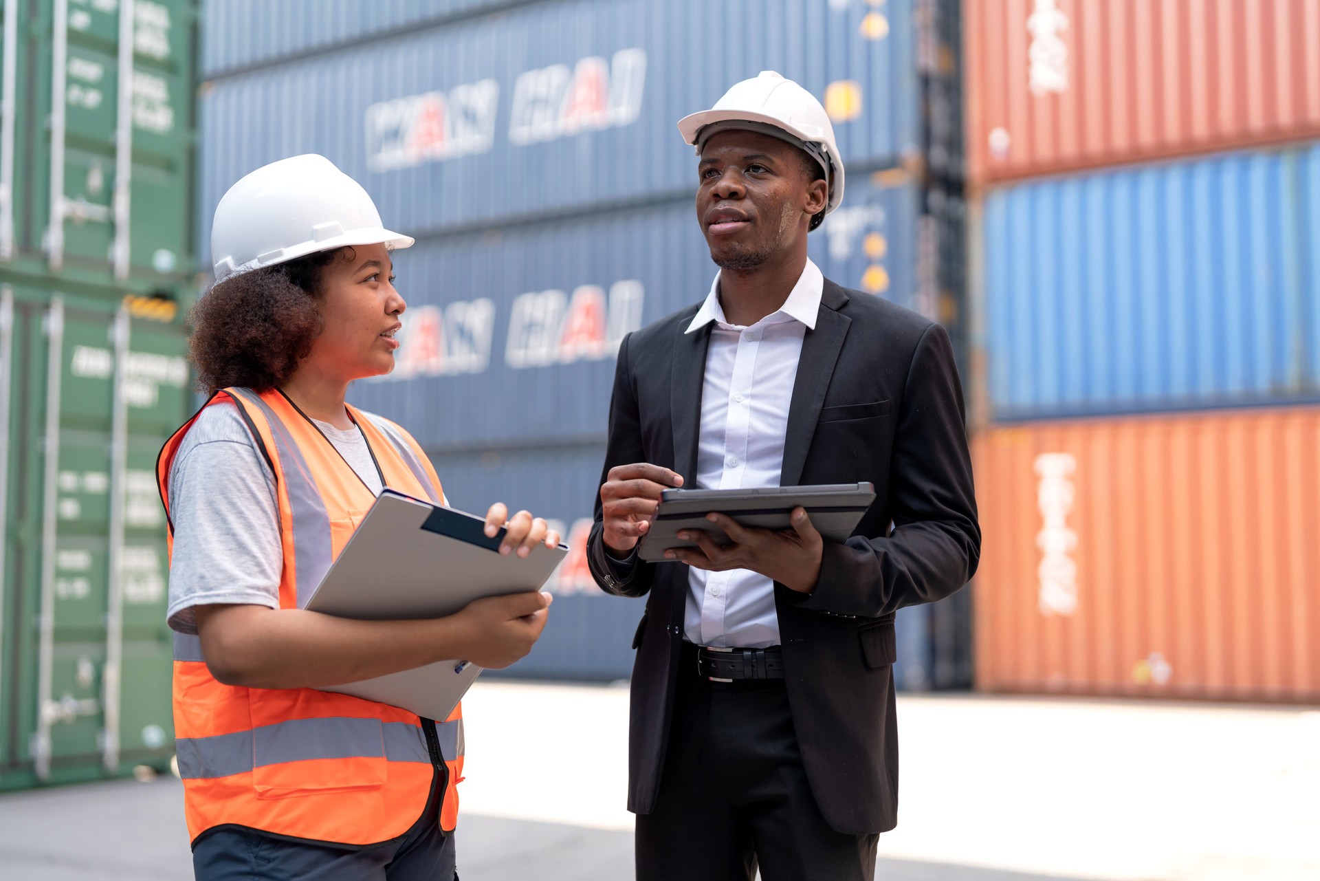 Black African dock manager standing and discussed over a tablet with engineer worker on the shipment in front of container box in the container warehouse area. Shipping Cargo Freights with Cargo Containers.